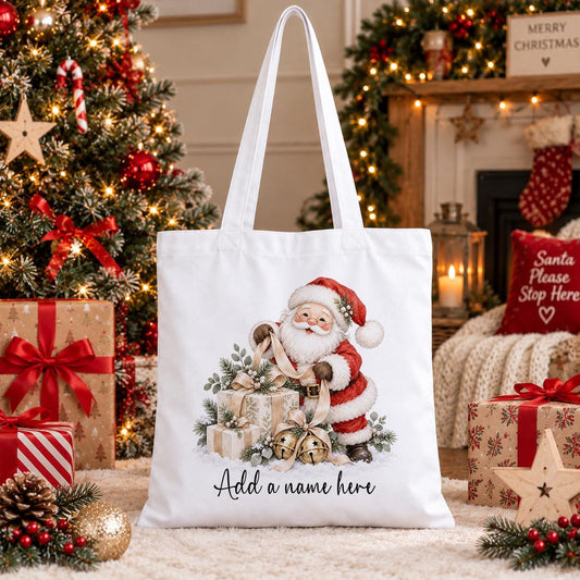 White cotton tote bag printed with Father Christmas reading his list beside wrapped presents and gold bells, with a personalised name below