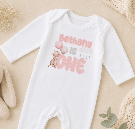 A white onesie with the text "Bethany is one" printed on it, along with a giraffe and balloons.