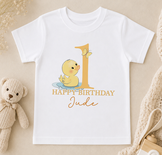 A white t-shirt with a cartoon duck and the text "Happy Birthday Jude" printed on it, along with a teddy bear and other baby items in the background.