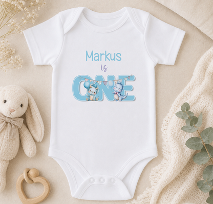White baby onesie printed with two cartoon dinosaurs either side of a bold 'ONE', personalised with the text 'Markus is ONE'.