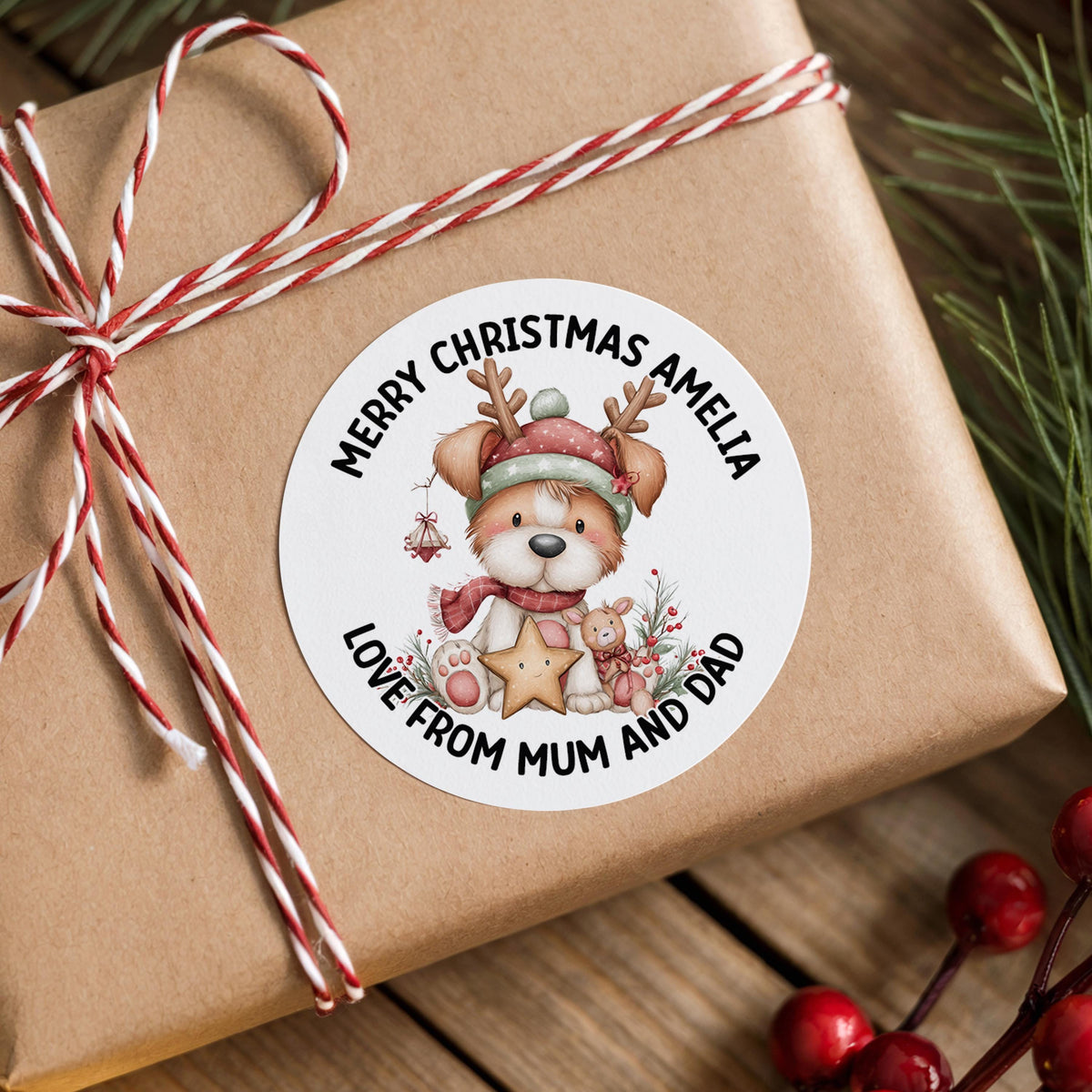 Round Christmas gift sticker featuring a cartoon reindeer dog wearing a Santa hat and scarf, surrounded by festive berries, personalised with a custom name, on a brown gift box