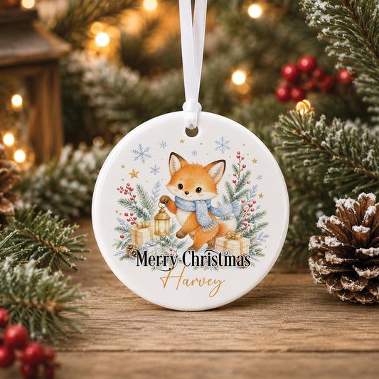 Personalised fox Christmas bauble, white ceramic ornament with a woodland fox, lantern and blue scarf, printed with a name and Merry Christmas, hanging on a Christmas tree