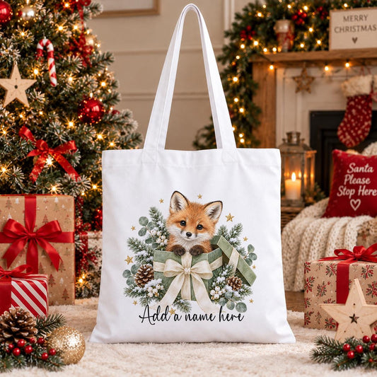 White cotton tote bag printed with a fox cub in a sage green gift box with pinecones, eucalyptus and gold stars, and a personalised name below