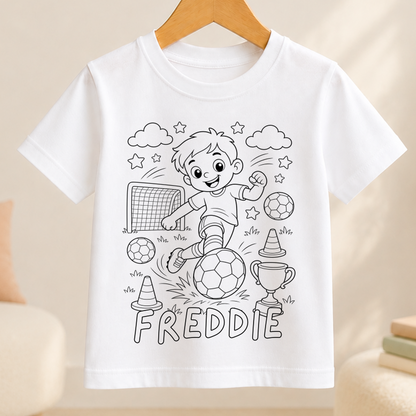 Line-art colour-in design of a boy kicking a football towards a goal, with a trophy and traffic cones, printed on a white kids' t-shirt with a personalised name below