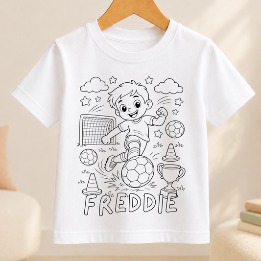 Line-art colour-in design of a boy kicking a football towards a goal, with a trophy and traffic cones, printed on a white kids' t-shirt with a personalised name below