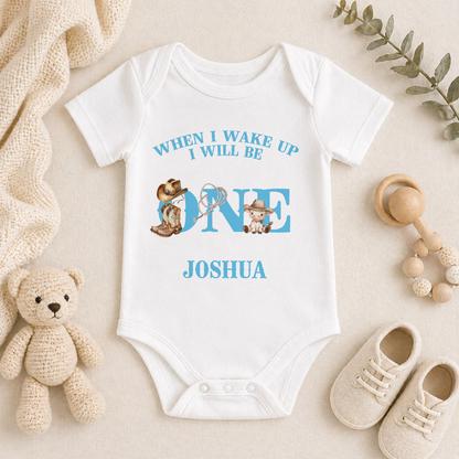 A white onesie with a personalized message, a teddy bear, baby shoes, and a wooden bead rattle are arranged on a beige surface.