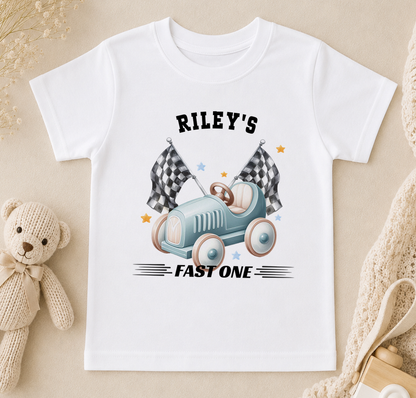 Personalised white t-shirt printed with a blue toy racing car, chequered flags and 'Riley's FAST ONE'.