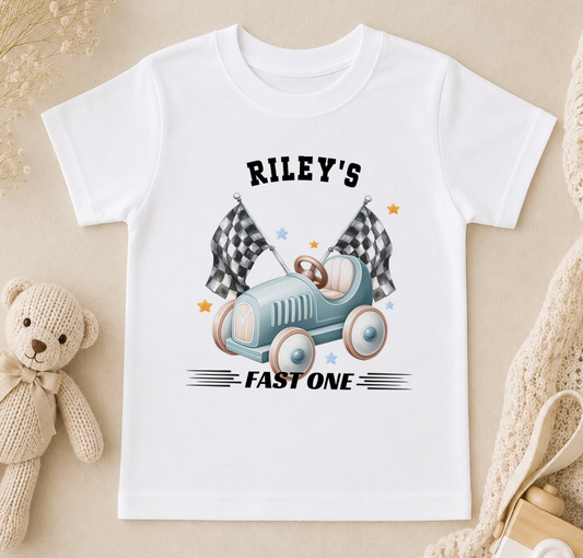 Personalised white t-shirt printed with a blue toy racing car, chequered flags and 'Riley's FAST ONE'.