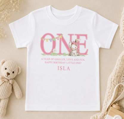 A white t-shirt with a pink text design that says "ONE" and features a small rabbit character. Below the text, there is a message that reads "A year of giggles, love and fun, happy birthday little one! ISLA".