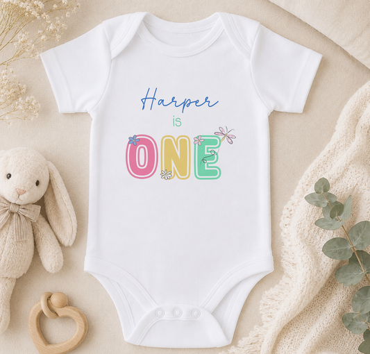 A white baby onesie with the text "Harper is ONE" printed on it, along with colorful floral designs. The onesie is placed on a beige surface, surrounded by a stuffed animal, a baby rattle, and some greenery.