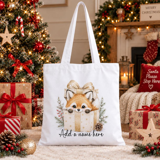 White cotton tote bag printed with a baby fox peeking out of a cream and gold gift box with pine sprigs and red berries, and a personalised name below