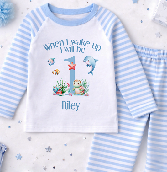 A white and blue striped pajama set with a personalized text and illustrations on the front, along with a blue pom pom and some small star-shaped decorations.