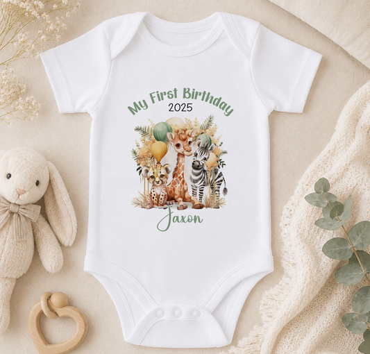 A white baby onesie with a printed design of animals and a balloon, along with the text "My First Birthday 2020/25" and "Jaxon" printed on it. The onesie is placed on a beige surface, surrounded by a stuffed animal, a wooden toy, and some greenery.