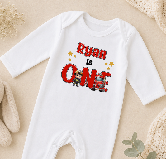 A white onesie with a red and black design, featuring the text "Ryan is one" and a cartoon character, is displayed on a beige surface.