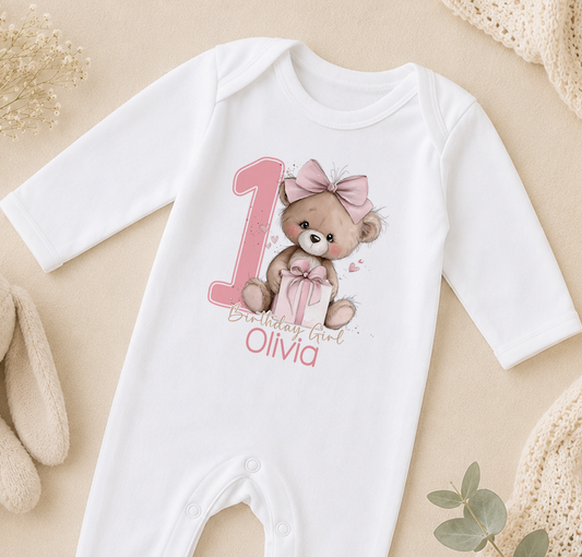A white onesie with a teddy bear and the number 1 printed on it, along with the text "Olivia" below the number.