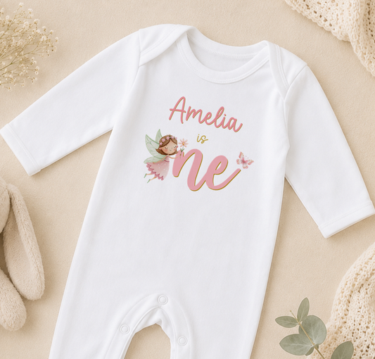 A white onesie with a personalized name and a fairy design, placed on a beige surface with some baby items and a plant in the background.