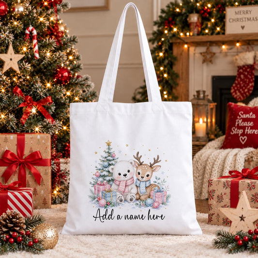 White cotton tote bag printed with a watercolour polar bear and baby reindeer in pastel scarves beside a Christmas tree, with a personalised name below