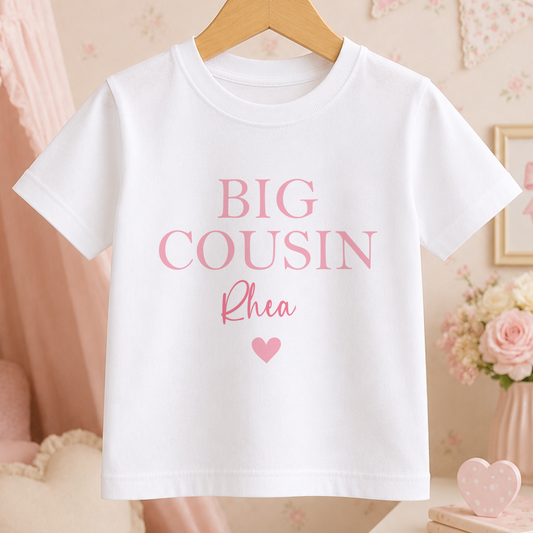 A white t-shirt with the text "BIG COUSIN Rhea" printed on it, along with a heart symbol.