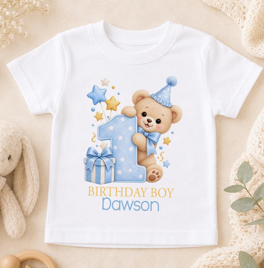 White baby 1st birthday top with a teddy bear in a blue party hat hugging a polka dot number one, and Birthday Boy above the name Dawson