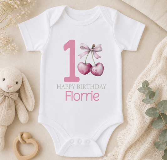 Personalised heart cherry and bow first birthday vest styled with a soft toy and greenery on a beige surface