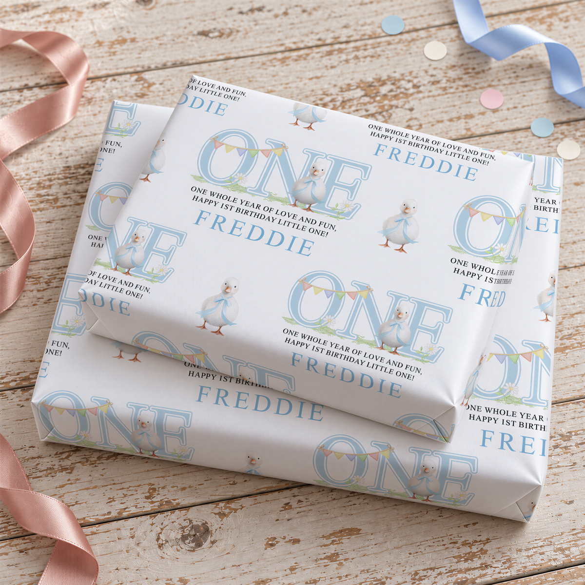 a set of wrapped presents with a repeating pattern of the name "FREDDIE" and a duck illustration. The presents are placed on a wooden surface, and there are some pink ribbons visible in the background.
