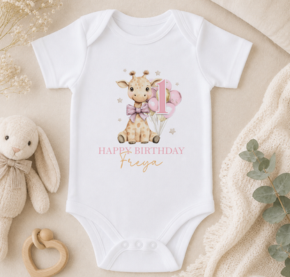 A white onesie with a giraffe and balloons, along with the text "Happy Birthday Freya", is displayed on a beige surface.