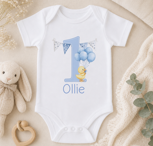 White baby onesie printed with a blue number '1' and yellow duckling holding balloon strings, styled with a stuffed animal and blanket.