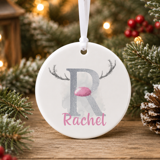 A white oval ornament with a silver letter "R" and a pink nose, hanging on a Christmas tree.