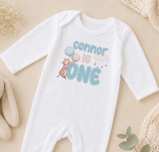 A white onesie with the text "Connor is one" printed on it, along with a giraffe and balloons.
