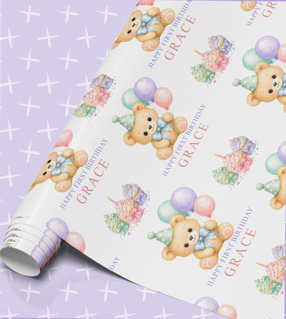 First birthday wrapping paper printed with watercolour teddy bears in party hats, pastel balloons, cupcakes and wrapped parcels, with Happy First Birthday and the name Grace in pink script on a white background, shown part unrolled
