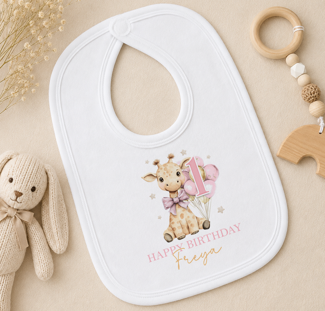 A white baby bib with a giraffe and balloons, along with a wooden toy and a wooden bead ring, are arranged on a beige surface.