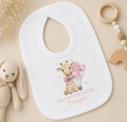 A white baby bib with a giraffe and balloons, along with a wooden toy and a wooden bead ring, are arranged on a beige surface.