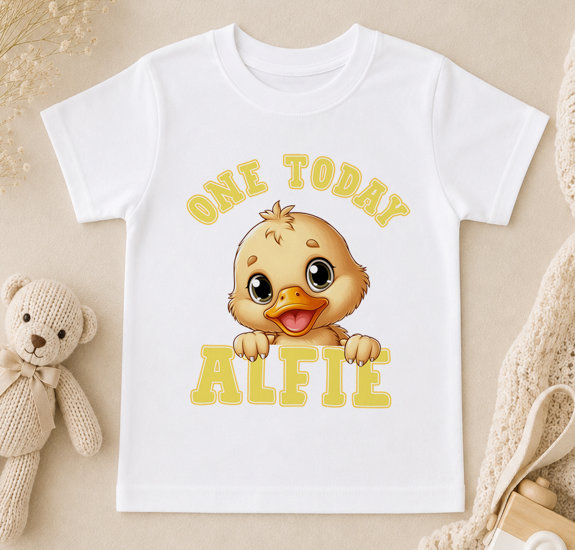 A white t-shirt with a cartoon duck and the text "ONE TODAY ALFIE" printed on it, along with a teddy bear and other baby items in the background.