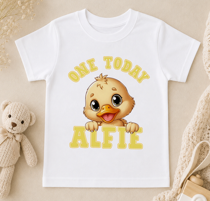 A white t-shirt with a cartoon duck and the text "ONE TODAY ALFIE" printed on it, along with a teddy bear and other baby items in the background.