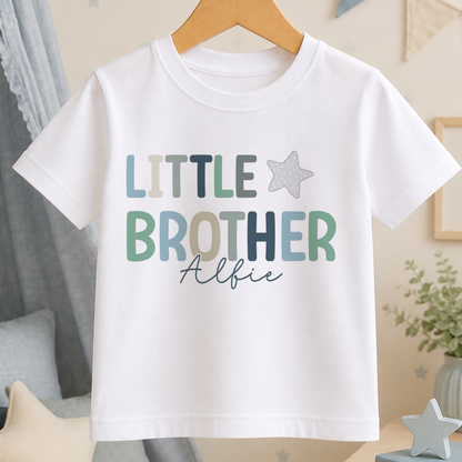 A white t-shirt with the text "LITTLE BROTHER" and a star graphic printed on it, hanging on a wooden hanger.