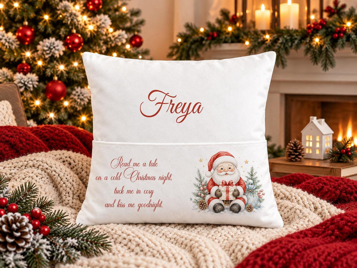 Personalised white Christmas book pocket cushion with Santa and presents and child’s name in red script, on a festive sofa