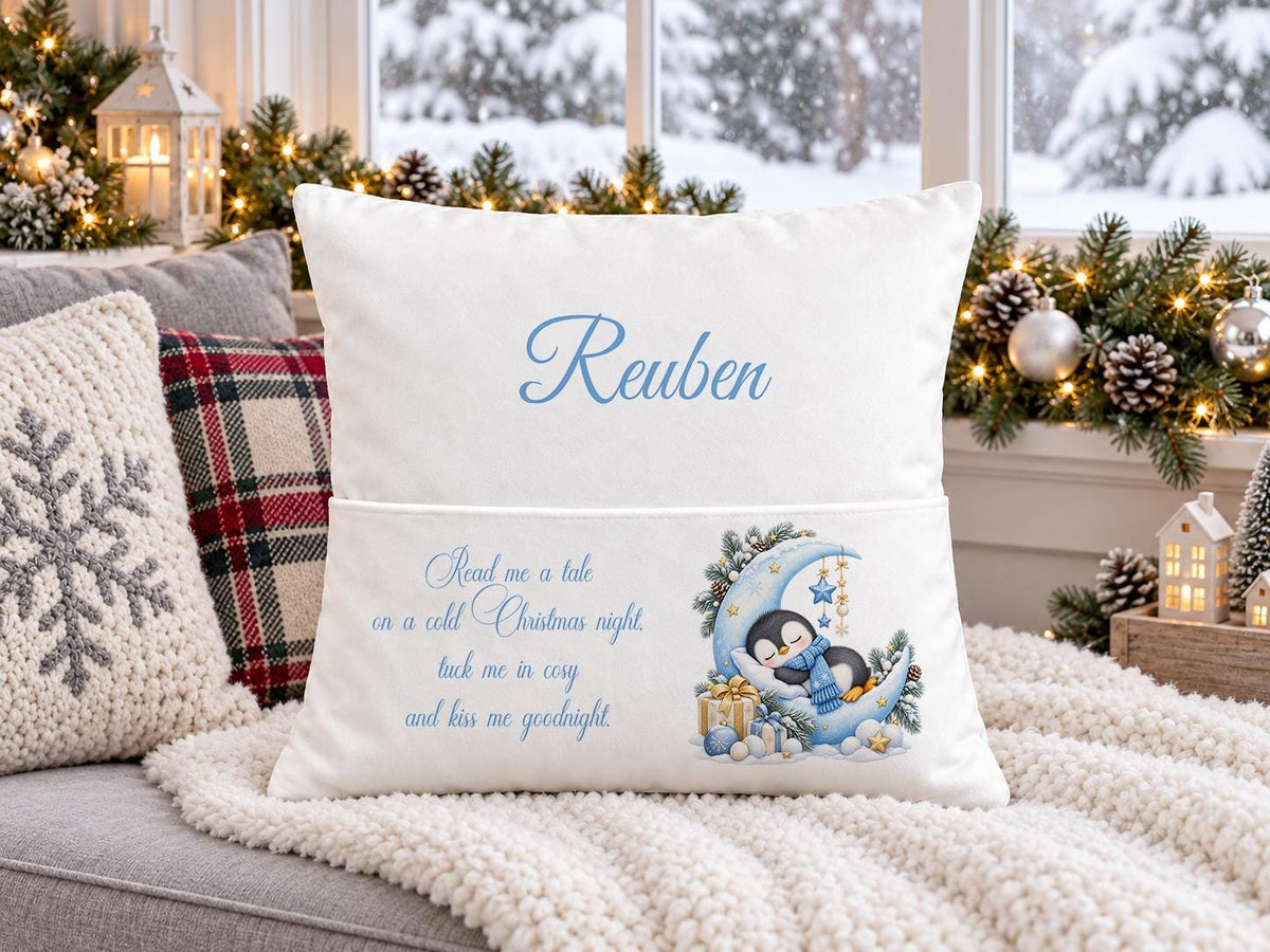 Personalised white Christmas book pocket cushion with a sleepy penguin on a crescent moon and child’s name in blue, on a festive sofa