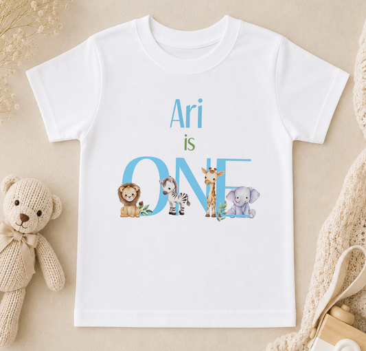 A white t-shirt with the text "Ari is ONE" printed on it, surrounded by illustrations of various animals.