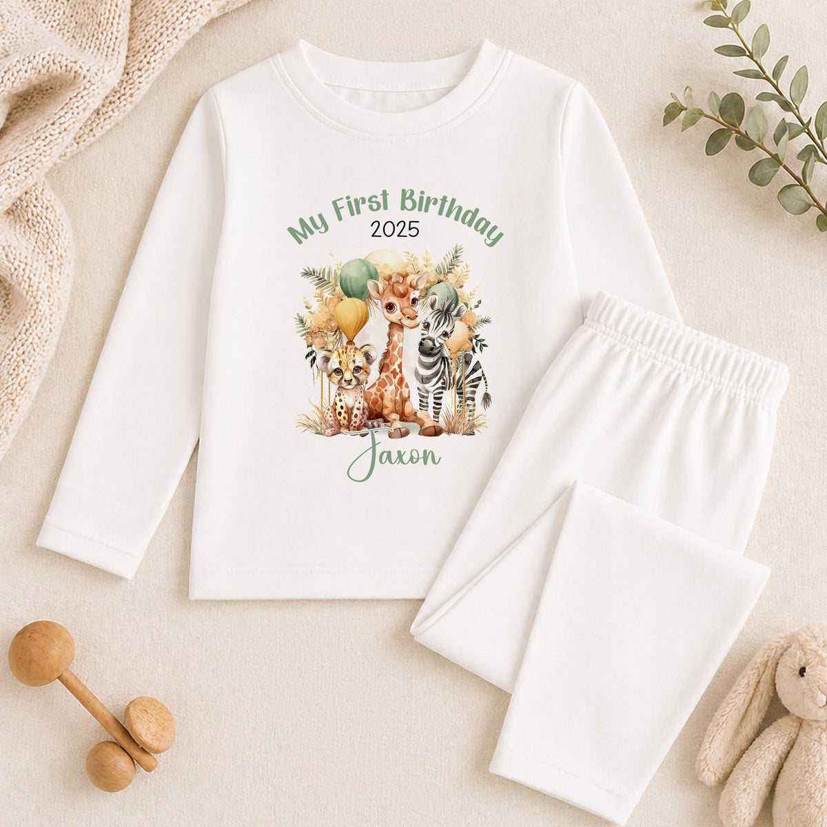 A white long-sleeved shirt with a printed graphic of a giraffe, zebra, and other animals, along with the text "My First Birthday 2025" and the name "Jaxon". The shirt is placed on a beige surface, and there are some stuffed animals and a wooden toy nearby.