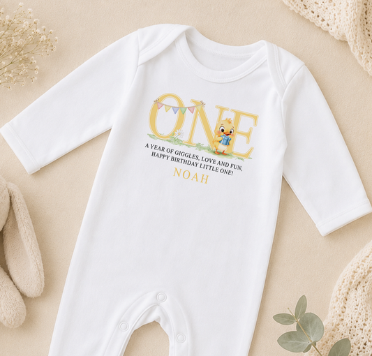 White baby onesie printed with a cartoon duck first birthday design and a personalised message, styled with keepsake props.