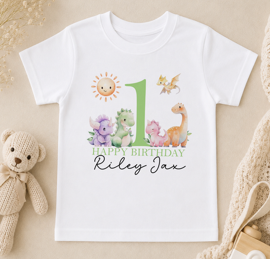 A white t-shirt with a birthday theme, featuring a green number one, a smiling sun, and various cartoon animal characters. The text "Happy Birthday Riley" is written below the image.
