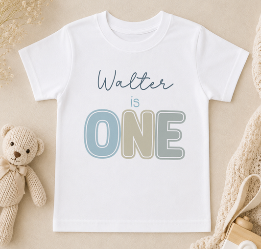 A white t-shirt with the text "Walter is ONE" printed on it, along with a teddy bear and some other baby items in the background.