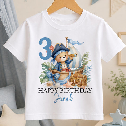 White cotton t-shirt printed with a teddy bear pirate in a boat with a treasure chest, a large age number, Happy Birthday and a name below.