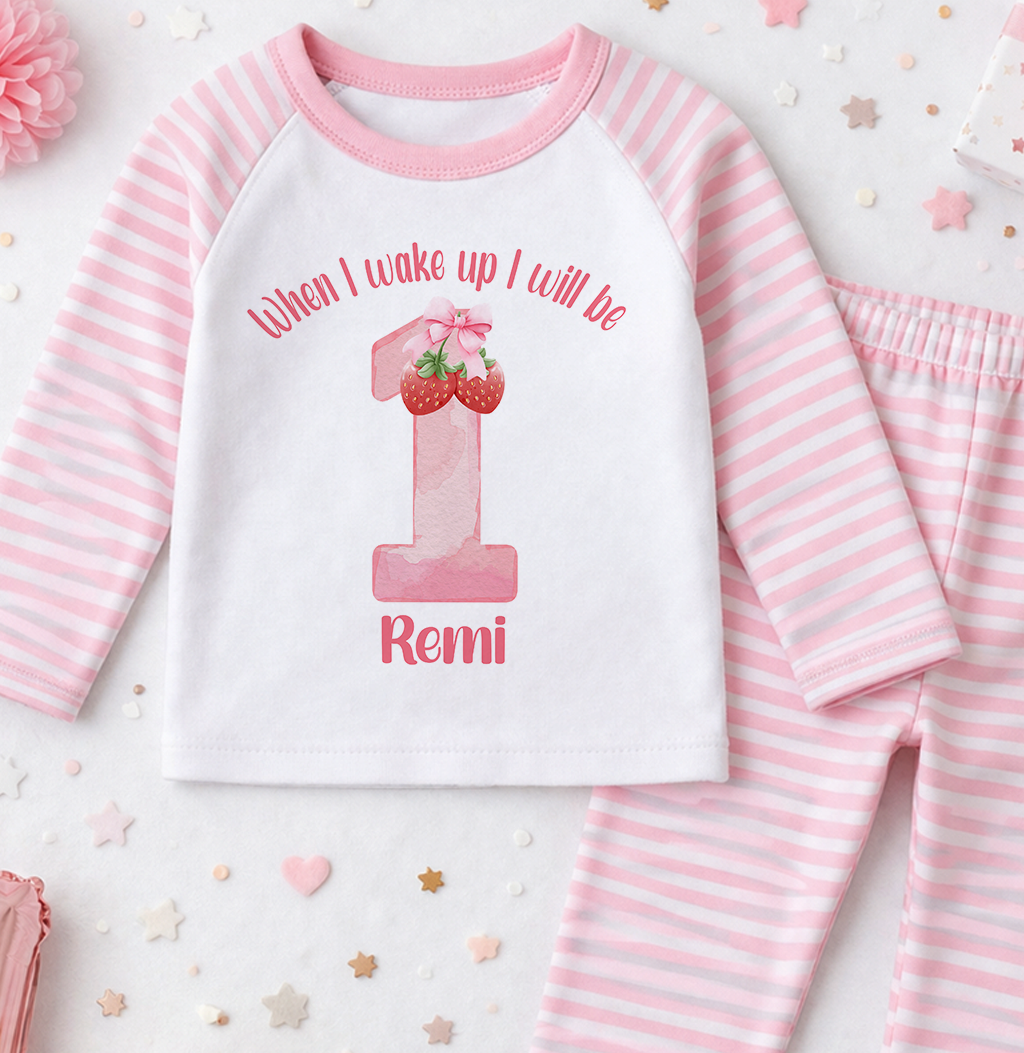 A pink and white striped onesie with a number 1 and strawberries printed on it, along with pink pajama bottoms.