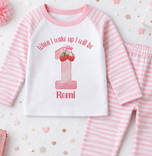 A pink and white striped onesie with a number 1 and strawberries printed on it, along with pink pajama bottoms.