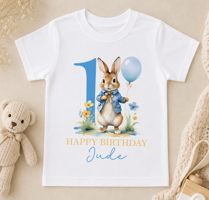 A white t-shirt with a graphic design featuring a rabbit holding a blue balloon and surrounded by flowers, with the text "Happy Birthday Jude" below the image.