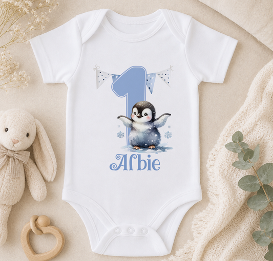 White baby onesie printed with a penguin design, a number 1 and the personalised name 'Alfie', laid out on a bed with a soft toy.
