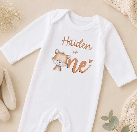 A white onesie with a cartoon fox and the text "Haiden is one" printed on it, along with a heart symbol.