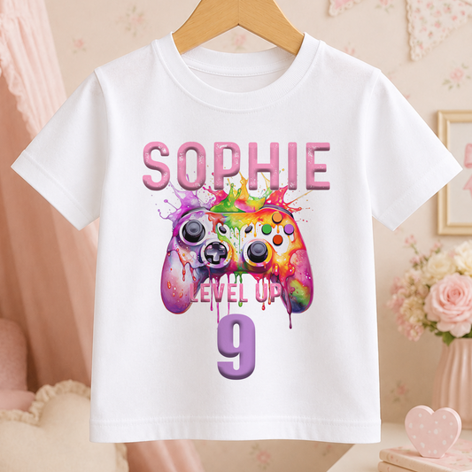 White cotton t-shirt with a colourful gaming controller design, pastel paint splats, and 'Level Up' text, personalised with a child's name and age