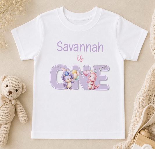 Personalised white t-shirt printed with a pink and a blue pastel dinosaur and 'Savannah is ONE' in purple.
